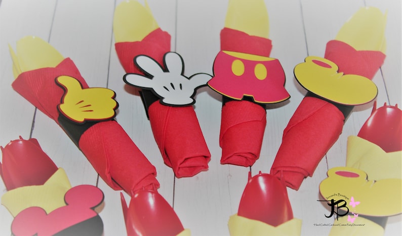 May include: Red cloth napkins with yellow utensils are decorated with Mickey Mouse-themed cutouts. The cutouts include a white glove, yellow thumbs up, red shorts, and a yellow shoe. The napkins are arranged on a white surface.