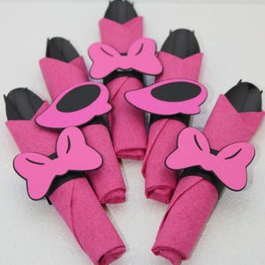 Op de afbeelding: Een verzameling roze servetten, elk opgerold en versierd met een zwarte en roze strik of een schoenvormige servetring. De servetten zijn in een waaiervorm gerangschikt, wat een decoratieve presentatie cre&euml;ert.