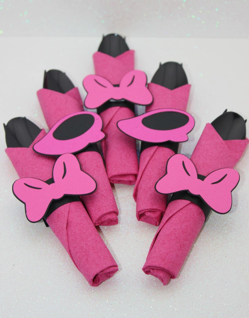 Op de afbeelding: Een verzameling roze servetten, elk opgerold en versierd met een zwarte en roze strik of een schoenvormige servetring. De servetten zijn in een waaiervorm gerangschikt, wat een decoratieve presentatie cre&euml;ert.