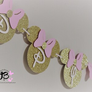 May include: A gold glitter Minnie Mouse themed banner with pink bows and white Disney lettering. The banner is strung together with white thread.