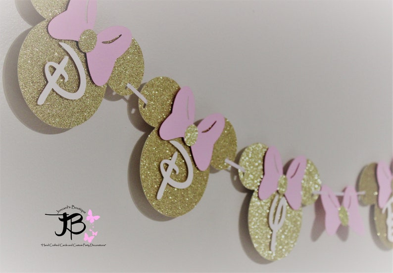 May include: A gold glitter Minnie Mouse themed banner with pink bows and white Disney lettering. The banner is strung together with white thread.