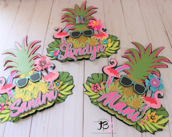 May include: Three tropical themed cake toppers featuring pineapples, flamingos, and glitter. The toppers are decorated with the names "Londyn", "Sandra", and "Alani".