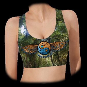 May include: A racerback sports bra featuring a forest print with green trees and foliage. Centered on the chest is a circular logo with orange and blue wings, containing the text Jamie Killen Art in a stylized, swirling blue and orange font.