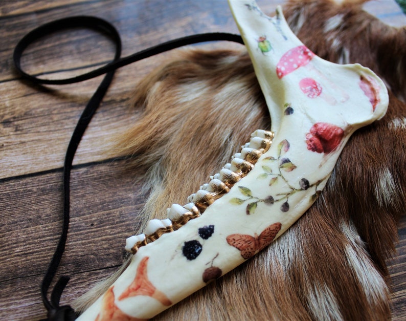 May include: A decorated animal jawbone with a black leather cord. The bone is painted with red mushrooms, butterflies, and berries. The teeth are painted gold. The jawbone rests on a brown and tan fur surface.