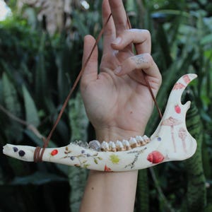 May include: A cream-colored jawbone pendant with painted botanical and mushroom designs, including strawberries and snails. The pendant is suspended by a brown leather cord. The jawbone features visible teeth and is decorated with small crystals.