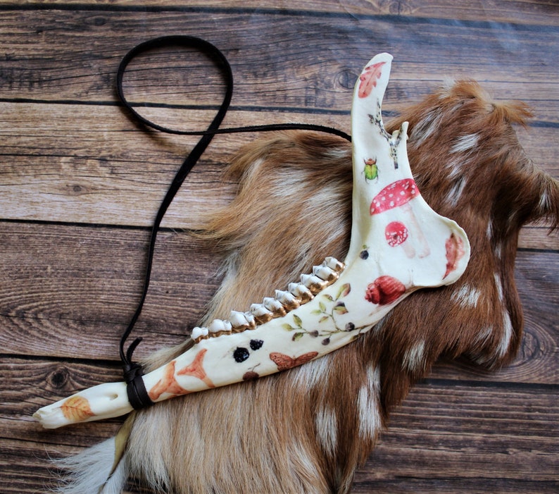 May include: A decorated animal jawbone with a black cord. The bone is painted with colorful illustrations of mushrooms, leaves, and insects. The jawbone rests on a brown and white fur pelt against a wooden background.