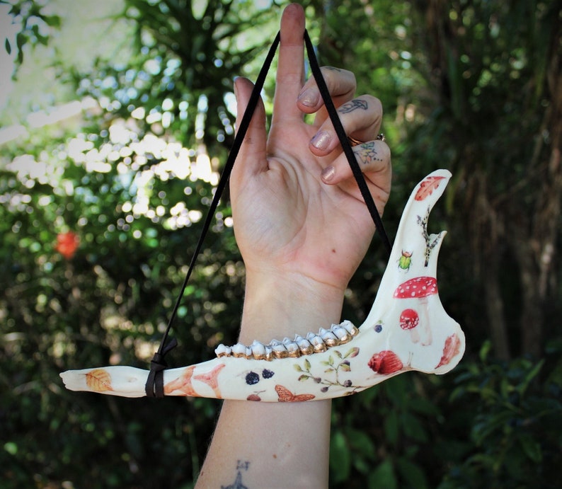 May include: A decorative bone jaw with a black hanging cord. The bone is painted with colorful illustrations of mushrooms, insects, and foliage. The teeth are accented with gold paint, creating a unique and artistic piece.