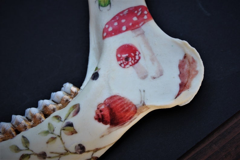 May include: A decorated animal jawbone featuring painted red mushrooms with white spots, a red snail, and gold-colored teeth. The bone is painted with green leaves and berries, set against a dark background.