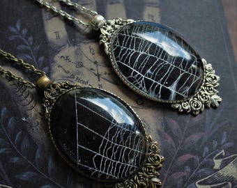 Puede incluir: Dos collares de plata y bronce con colgantes ovalados que presentan un diseño de telaraña en blanco y negro. Los colgantes están rodeados de acentos decorativos en forma de rosa.