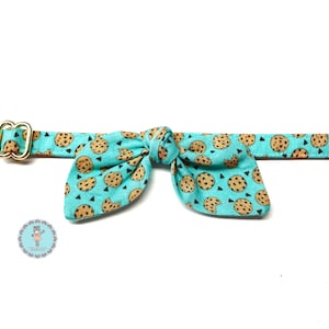 May include: A turquoise pet collar with a bow tie, featuring a pattern of chocolate chip cookies and black triangles. The collar has a gold-colored metal buckle and a matching bow tie.