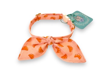 May include: A pink pet collar with a bow tie. The collar and bow tie are patterned with orange croissants. The collar has a gold-colored clasp and a tag with a cartoon illustration.