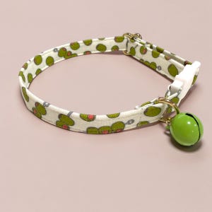 May include: A pet collar featuring a repeating pattern of green olives with red pimentos on a cream-colored fabric background. The collar includes a white plastic side-release buckle, a gold-toned metal D-ring, and a round, bright green metal bell.