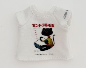 May include: White doll-sized t-shirt with a graphic of a black cat reading a book. The cat is wearing a yellow shirt and red pants. Japanese text is printed above the cat.