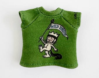 May include: Green t-shirt featuring a cartoon character holding a flag that reads "WILD FOREVER." The character is wearing a crown and has a black tail. The t-shirt is a solid green color.