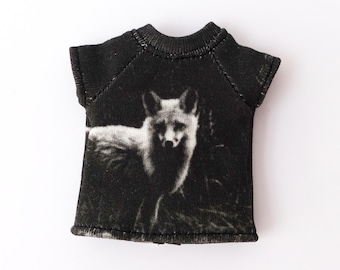 May include: Black t-shirt featuring a grayscale image of a fox. The fox is standing in a grassy area. The t-shirt has short sleeves and a crew neck. The image is centered on the front of the shirt.