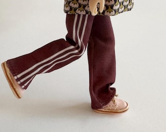 May include: A miniature doll dressed in burgundy track pants with white stripes and a patterned jacket. The doll is wearing light pink shoes with tan soles. The jacket has a floral pattern in shades of green, pink, and black.