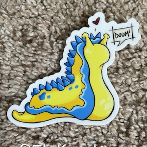 May include: A yellow and blue sticker featuring a cartoon sea slug with the word "DOOM!" in a speech bubble. A small red heart is above the speech bubble. The sticker has the text "@ElyKreations" at the bottom.