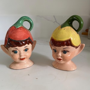 May include: A pair of ceramic salt and pepper shakers. One shaker is red and green, the other is yellow and green. Both shakers are shaped like a face with a flower-like top and a handle. The faces have blue eyes and red lips.