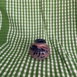 Può includere: Un piccolo vaso rotondo in ceramica con uno schema di colori rosa e blu. Il vaso ha un bordo blu scuro e presenta un motivo floreale blu scuro. Lo sfondo è un motivo a scacchi verdi e bianchi.