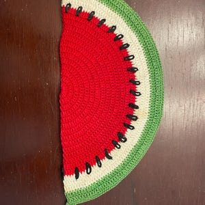 Puede incluir: Un artículo de ganchillo en forma de media sandía con un centro rojo, un medio blanco y una corteza verde. Las puntadas negras representan semillas. Un lazo verde está unido en la parte inferior. Este artículo hecho a mano es probablemente un accesorio de cocina, como un salvamanteles.