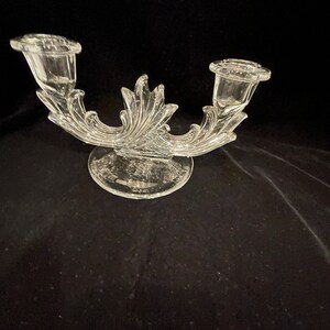 May include: A clear glass candle holder with two arms and a decorative base. The arms are shaped like leaves and the base is round with a raised design.