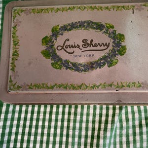 Könnte beinhalten: Vintage rechteckige Metallschale mit verblasster rosa Oberfläche. Die Schale trägt die Aufschrift "Louis Sherry New York" in eleganter Schrift, umgeben von einem floralen Kranz. Der Rand ist mit grünen und weißen floralen Akzenten verziert.