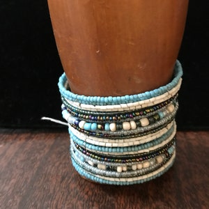 May include: A stack of beaded bracelets in shades of blue, white, and black. The bracelets are made of small, round beads and are layered on top of each other.