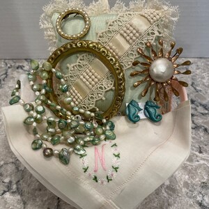 Pode incluir: Um travesseiro de renda branca com uma moldura dourada, um colar de pérolas, um broche dourado com um centro de pérola e um broche de cavalo-marinho azul. Os itens estão em um lenço de linho branco com um "N" bordado.