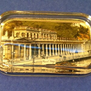 May include: A rectangular glass ashtray with a detailed image of a building with columns and a bridge. The image is in shades of brown, beige, and green. The text "Karl Vary" is visible on the image.