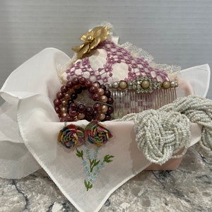 May include: A white handkerchief with embroidered flowers, a pink and white lace pillow, a brown and pink beaded bracelet, a white beaded necklace, a silver hair comb with rhinestones, and a gold flower brooch.