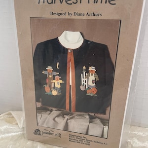 Może przedstawiać: Wzór do rękodzieła na czarny kardigan z motywem żniw. Wzór przedstawia dwie kukły z dyniami i płotem. Wzór nosi tytuł "Harvest Time" i został zaprojektowany przez Diane Arthurs. Wzór pochodzi z The Powder Mill.