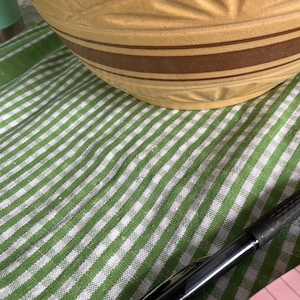Może przedstawiać: Duża, beżowa ceramiczna misa do mieszania z brązowymi paskami i detalami w kształcie liści. Misa stoi na zielono-białej, pasiastiej tkaninie. Czarny długopis spoczywa na tkaninie.
