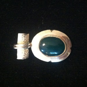 May include: Silver brooch featuring a dark teal oval stone. The brooch has a textured bar and a ring detail connecting to the oval setting. The setting has a textured border. The brooch is set against a black background.