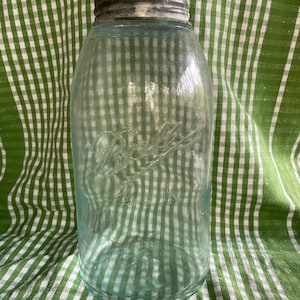 Puede incluir: Un tarro Mason Ball vintage teñido de azul con tapa metálica. El tarro tiene letras en relieve y un diseño clásico. El tarro se exhibe sobre una tela a cuadros verde y blanca.