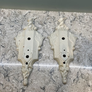 May include: Two antique white decorative wall sconces with ornate, scrollwork designs. Each sconce has a rectangular shape with three holes and a decorative top. The sconces are made of metal and have a distressed finish.