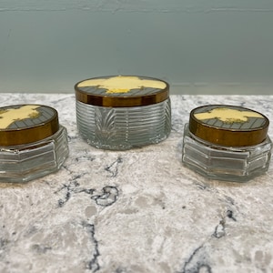 Könnte beinhalten: Set aus drei Vintage-Gläsern mit goldenen Deckeln und dekorativen Oberteilen. Die Gläser sind aus klarem Glas mit einem strukturierten Design. Die Deckel sind aus goldenem Metall mit einem weißen und goldenen Design.