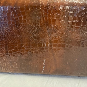 May include: A vintage brown suitcase with a textured, crocodile-like pattern. The suitcase has brass-colored hardware, including clasps and a handle. The surface shows signs of wear, adding to its antique charm.