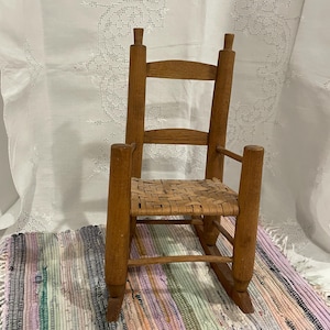 May include: A small wooden rocking chair with a woven seat. The chair is made of natural wood and has a rustic look. It is sitting on a colorful striped rug.