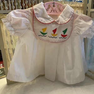 May include: White baby dress with a Peter Pan collar trimmed in red. The bib features embroidered sailboats in yellow, red, and green. The dress has short, puffed sleeves and is hanging on a pink hanger.
