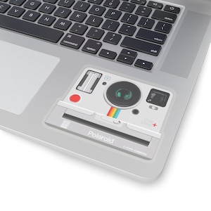 Könnte beinhalten: Ein silberner Laptop mit einem Aufkleber einer Polaroid OneStep+ Kamera. Der Aufkleber zeigt ein weißes Kameragehäuse mit schwarzem Objektiv, rotem Knopf und Regenbogenstreifen. Der Text "Polaroid" und "1-TYPE CAMERA" sind sichtbar.