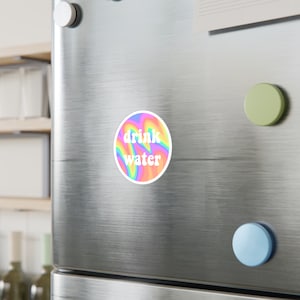 May include: A circular sticker featuring a rainbow swirl pattern and the words "drink water" in white. It's affixed to a stainless steel fridge door, alongside a white, green, and blue magnet.