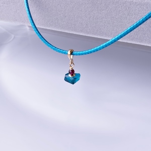 Puede incluir: Un collar de cordón azul con un pequeño colgante que presenta una piedra preciosa azul y una cuenta roja de acento.