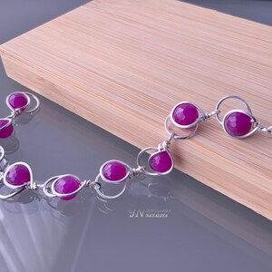 May include: A silver bracelet with a clasp fastening features a series of pink gemstone beads connected by silver wire loops. The bracelet is displayed on a wooden surface.