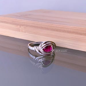 May include: A silver ring featuring a bright pink gemstone, set within a woven silver design. The ring sits on a reflective surface, with a wooden box in the background. The design of the ring is detailed and striking, showcasing a piece of jewellery.