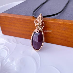 以下が含まれることがあります： 大きな楕円形の濃い紫色の宝石が付いた、金色のペンダント。宝石は金のワイヤーフレームに収められ、黒いコードから吊り下げられています。ペンダントは木製の箱の上に置かれています。「JN Natalia」の文字が見えます。