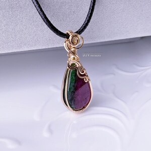 May include: A gold-coloured wire-wrapped pendant with a teardrop-shaped gemstone. The gemstone displays deep red and green hues. The pendant is suspended from a black cord. The text "JIV NATALIA" is visible on the pendant.