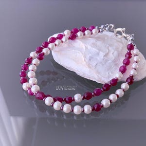 Może przedstawiać: Bransoletka z dwoma sznurami z naprzemiennymi koralikami w kolorze rubinu i białymi perłami. Biżuteria jest zabezpieczona srebrnym zapięciem. Bransoletka jest prezentowana na białej, teksturowanej powierzchni.
