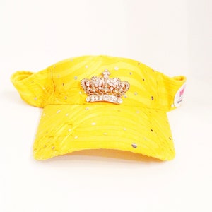 May include: A yellow visor with a rhinestone crown design. The visor has a small logo on the side.