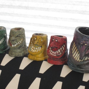 Puede incluir: Una colección de cinco tazas de cerámica con caras de monstruos. Las tazas están pintadas en varios colores, incluyendo verde, gris, amarillo, rojo y morado. Cada taza presenta una cara de monstruo sonriente única con dientes y rasgos faciales detallados.