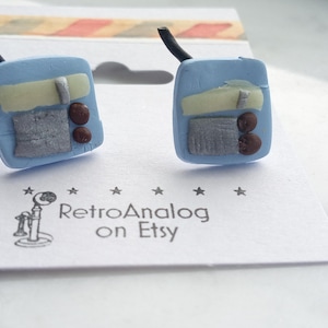 May include: Handmade blue earrings in the shape of retro mobile phones. Each earring has a black antenna, a light yellow rectangle, a grey rectangle, and two brown circles. The earrings are on a white surface with the text "RetroAnalog on Etsy".